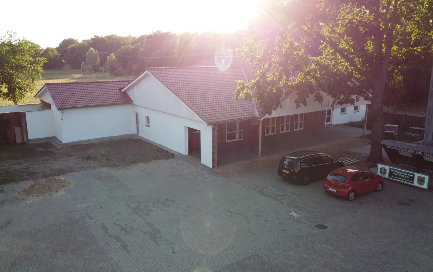 Das Dorfgemeinschaftshaus, ein weiß-rotes Gebäude mit Parkplatz davor, im Gegenlicht der tiefstehenden Sonne
