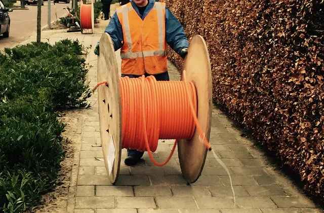 Ein Bauarbeiter in Warnweste zieht eine Kabeltrommel mit orangefarbenem Leerrohr über einen Gehweg