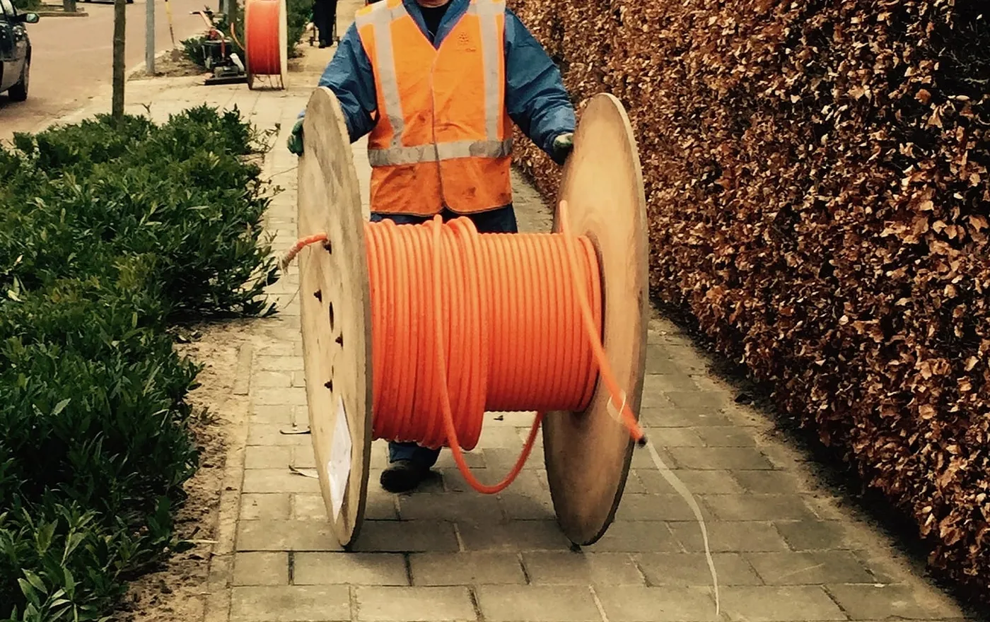 Ein Bauarbeiter in Warnweste zieht eine Kabeltrommel mit orangefarbenem Leerrohr über einen Gehweg
