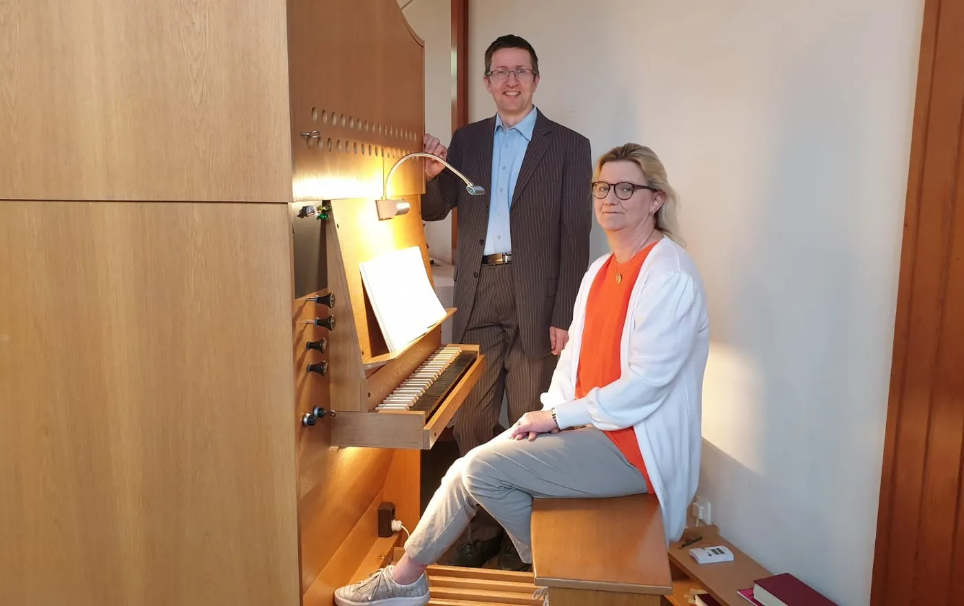 Antje Hartmann sitzt an der Orgel, neben ihr steht ein Mann im Anzug; beide blicken in die Kamera