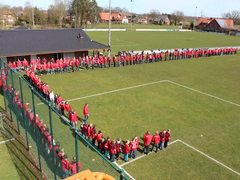 Luftbild des United Park: Hunderte Menschen in roten Trikots stehen in einer langen Reihe am Rand des Sportplatzes